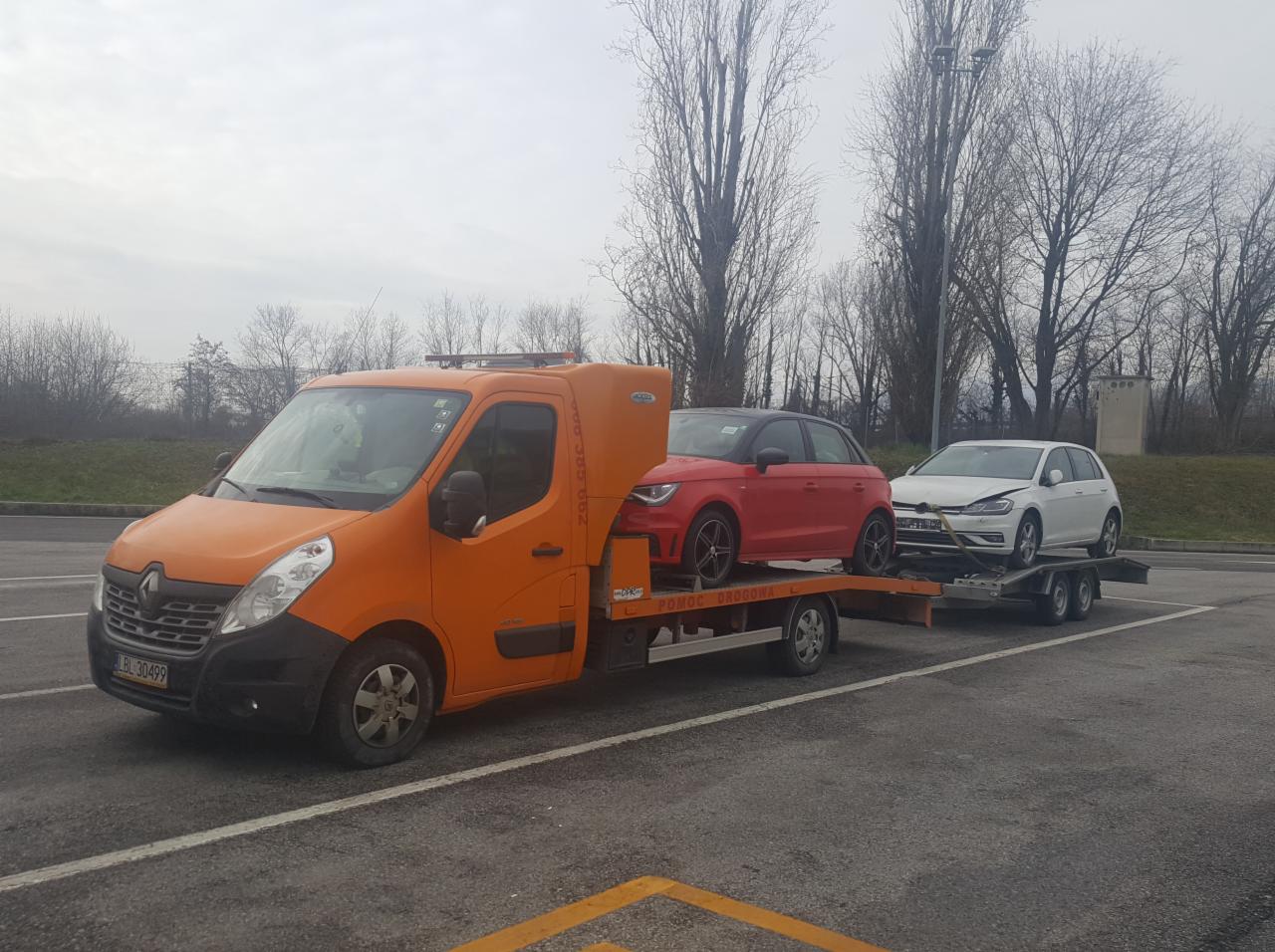 Pomarańczowa laweta Renault z dwoma samochodami osobowymi na przyczepie: czerwonym Audi A1 i uszkodzonym białym Volkswagenie Golfie, zaparkowana na parkingu z drzewami w tle.