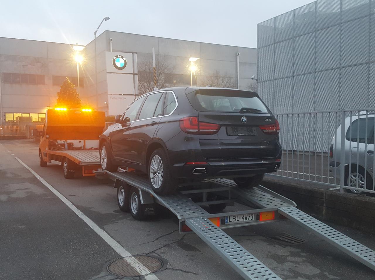 Czarny samochód BMW X5 na metalowej lawecie z pomarańczowym pojazdem holowniczym w tle, przed budynkiem z logo BMW.