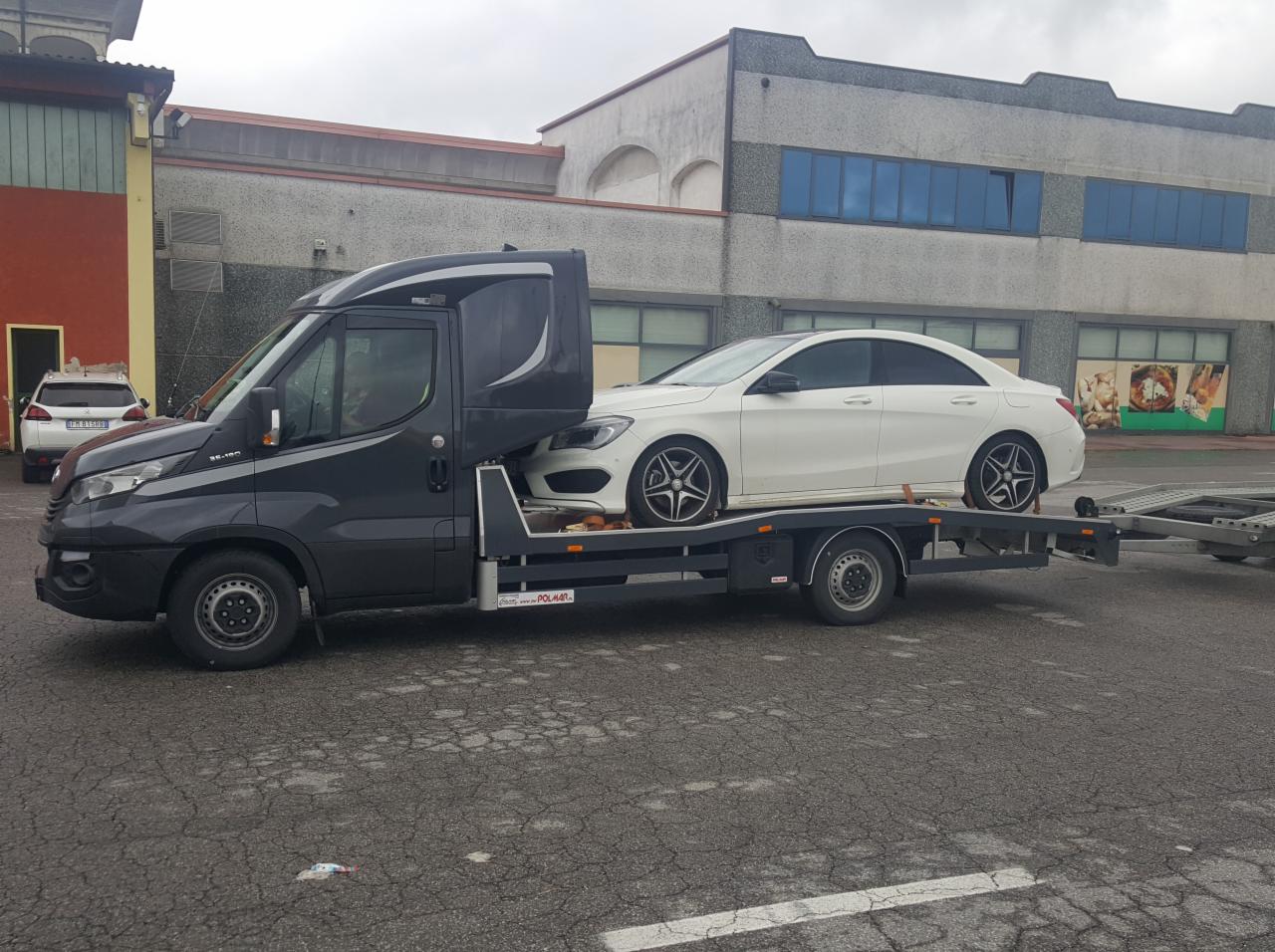Czarny Iveco Daily z platformą przewozi biały samochód osobowy Mercedes CLA na parkingu przed budynkiem.