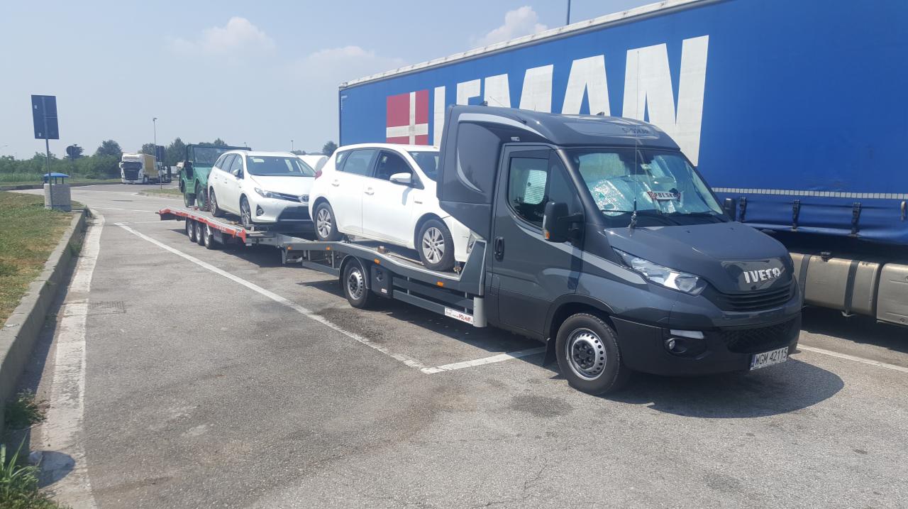 Szara laweta Iveco z dwoma białymi samochodami osobowymi na przyczepie, zaparkowana na parkingu z niebieską naczepą w tle.