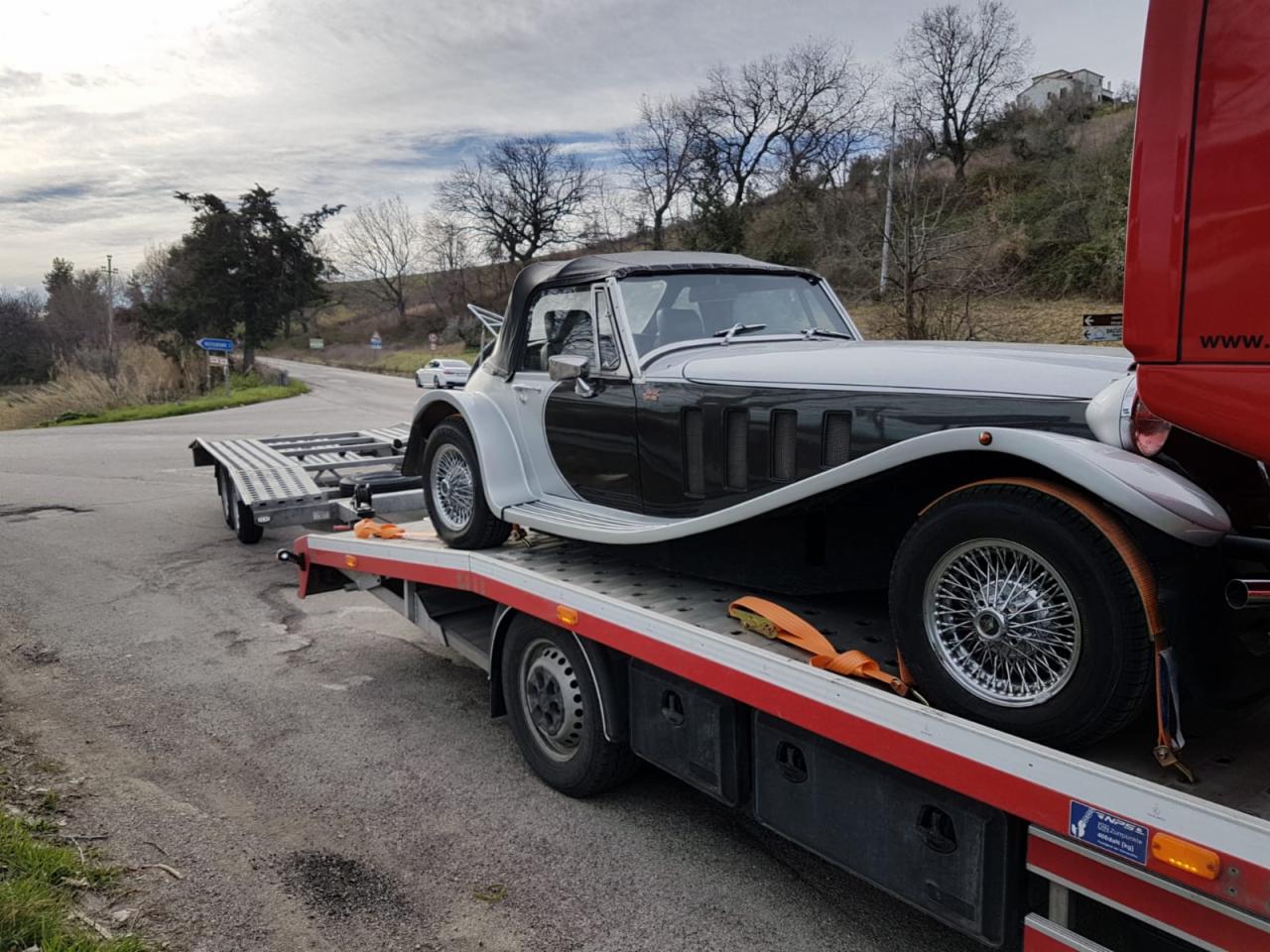 Czarno-biały zabytkowy kabriolet na lawecie z pomarańczowymi pasami zabezpieczającymi, w tle krajobraz z drzewami i budynkami.