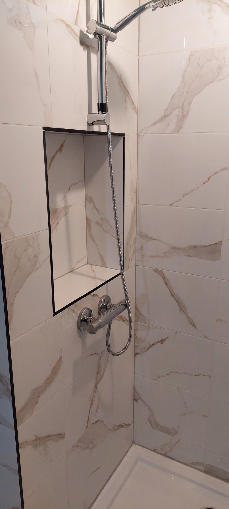 Nowoczesna kabina prysznicowa z chromowaną armaturą i wnęką wykończoną czarną ramą w płytkach imitujących marmur.