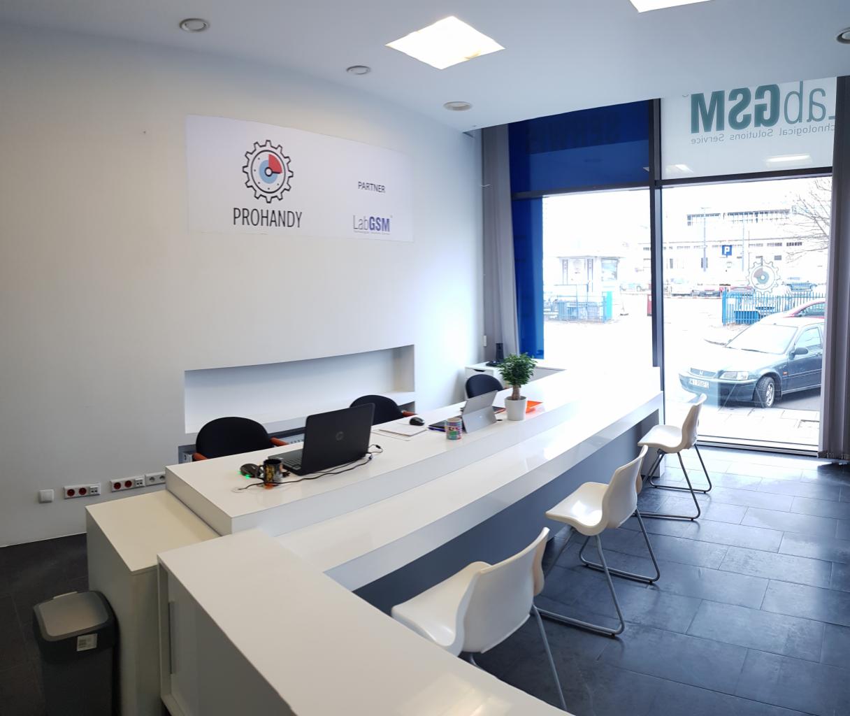 Jasne wnętrze biura serwisowego z długim białym blatem, laptopem, kubkiem z kolorowym nadrukiem i logotypami Prohandy i LabGSM na ścianie.