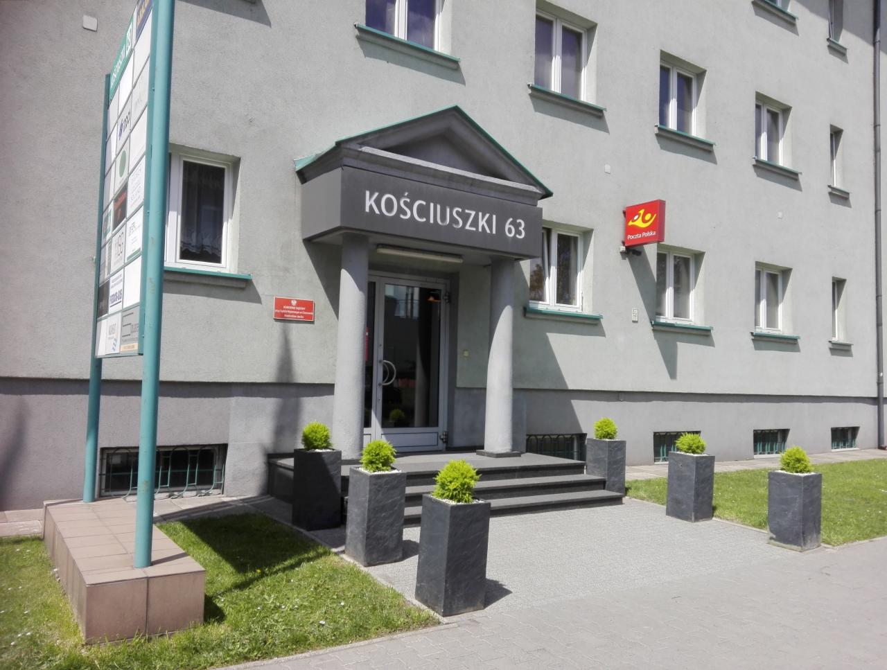 Szary budynek z napisem 'Kościuszki 63' nad wejściem, kolumnami i donicami z zielonymi krzewami, z logo Poczty Polskiej na ścianie.