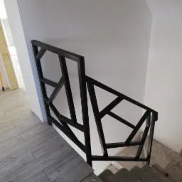 Czarna, geometryczna poręcz schodowa z metalu, widziana z góry, na tle białych ścian i szarych płytek podłogowych. Widoczne niedokończone wykończenie ścian.