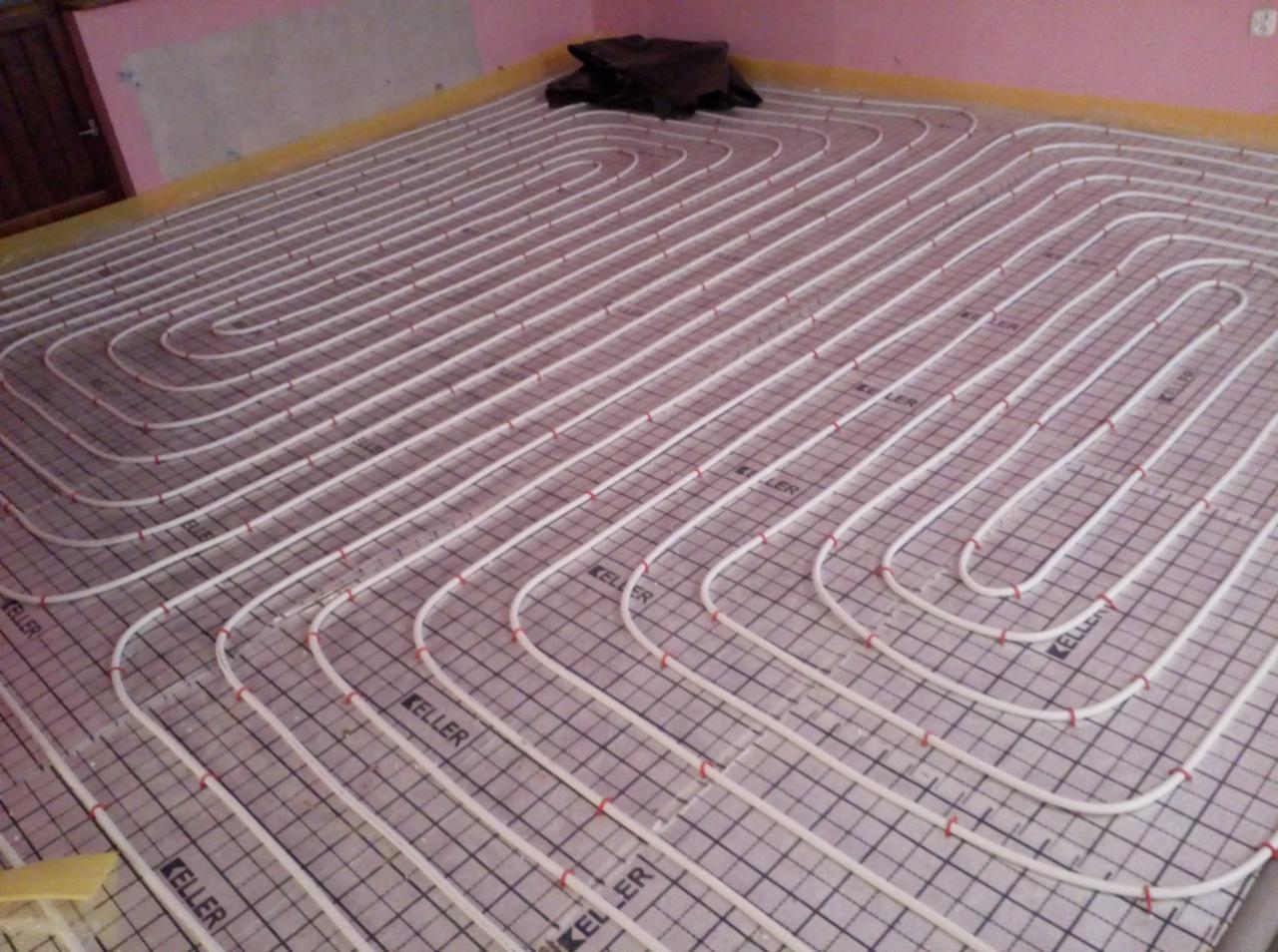 Białe rury ogrzewania podłogowego ułożone w pętle na siatce z oznaczeniami 'KELLER', przymocowane czerwonymi klipsami, widoczne w trakcie instalacji.