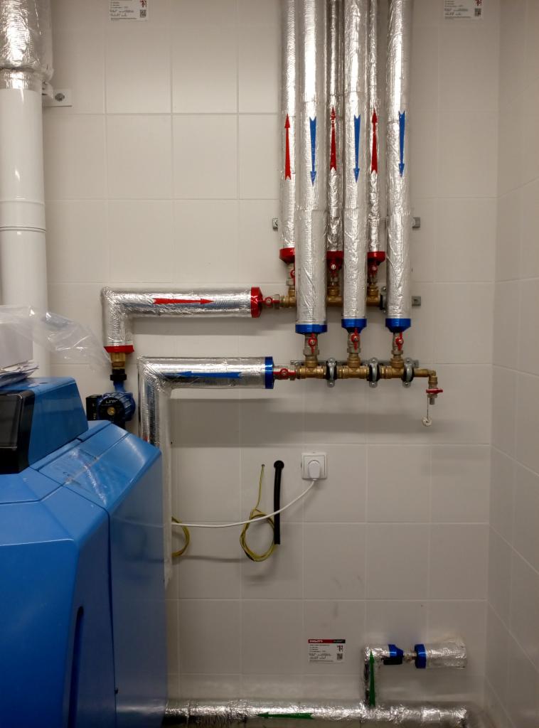 Instalacja grzewcza z kotłem, rurami w izolacji aluminiowej oznaczonymi strzałkami kierunku przepływu, zamontowana na białej, kafelkowej ścianie.