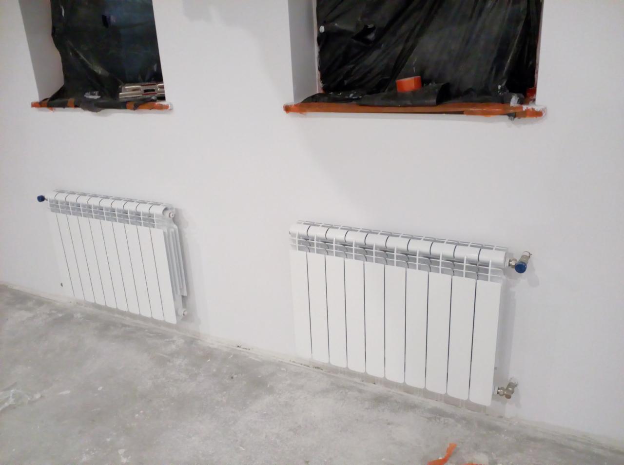 Dwa białe grzejniki aluminiowe zamontowane na ścianie w surowym stanie, okna zabezpieczone folią malarską, betonowa podłoga.