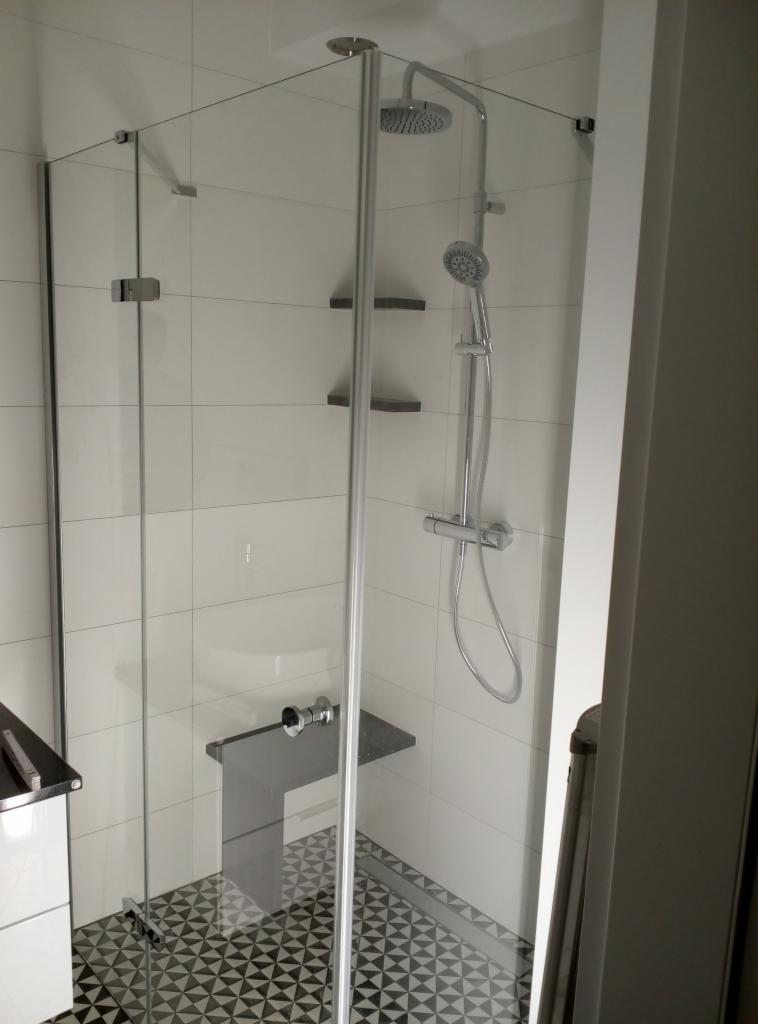 Nowoczesna kabina prysznicowa z deszczownicą i siedziskiem, wykończona białymi płytkami i geometryczną posadzką.