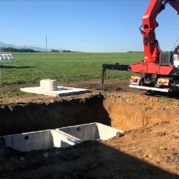 Ciężarówka z dźwigiem opuszcza betonowe elementy do głębokiego wykopu na tle zielonego pola z belami siana i pasmem górskim w oddali.