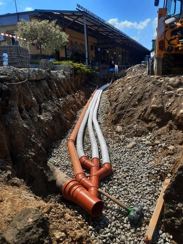 Wykop z ułożonymi rurami kanalizacyjnymi, zasypany drobnym żwirem, widoczny fragment koparki i budynek w tle.