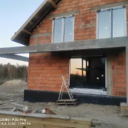 Surowy stan budowy domu jednorodzinnego z zamontowanymi oknami, widoczne cegły, betonowe nadproża, drabina oparta o ścianę, piasek i drewniane deski na pierwszym planie.