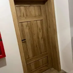 Zamknięte, drewniane drzwi wewnętrzne z panelem dekoracyjnym i czarnym, minimalistycznym uchwytem na tle białej ściany, widoczny fragment czerwonej skrzynki.