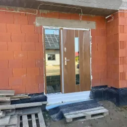 Nowo zamontowane drzwi wejściowe w brązowej okleinie, jeszcze z folią ochronną, w surowym murze z czerwonej cegły kratówki, z widocznymi elementami montażu i paletami budowlanymi u dołu.