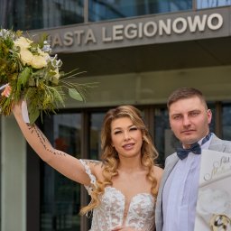 Studio Filmowe Videoefekt - Para młoda pozuje z bukietem i aktem małżeństwa na tle budynku z napisem MIASTA LEGIONOWO. Panna młoda z tatuażem na ramieniu.