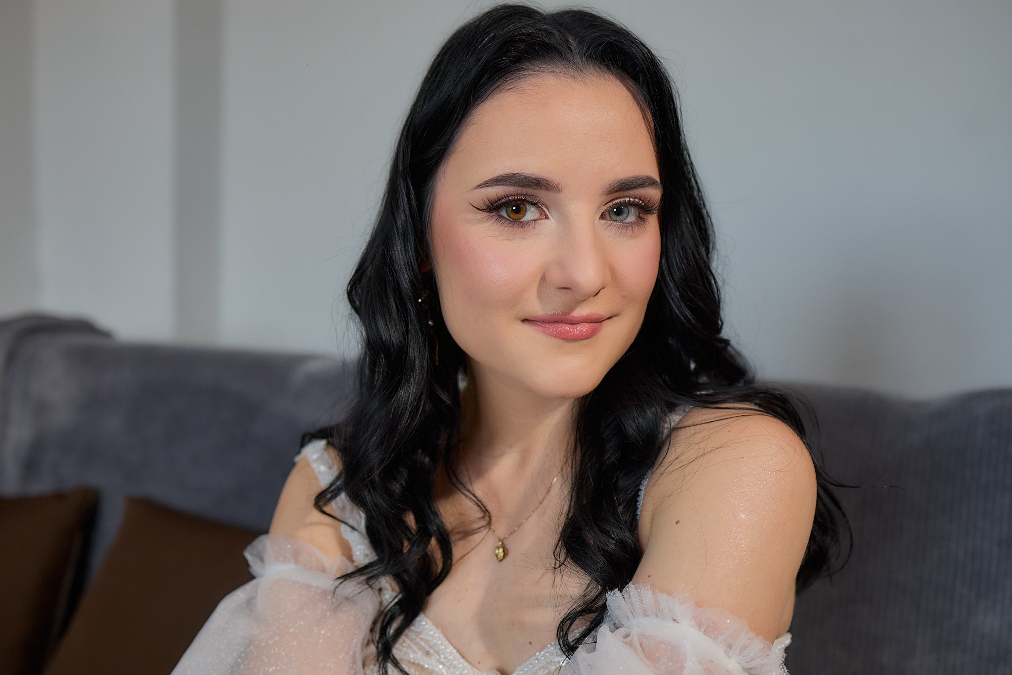 Portret uśmiechniętej kobiety o heterochromii, w białej sukni z falbankami, na tle szarej kanapy i ściany. Makijaż podkreśla urodę modelki.