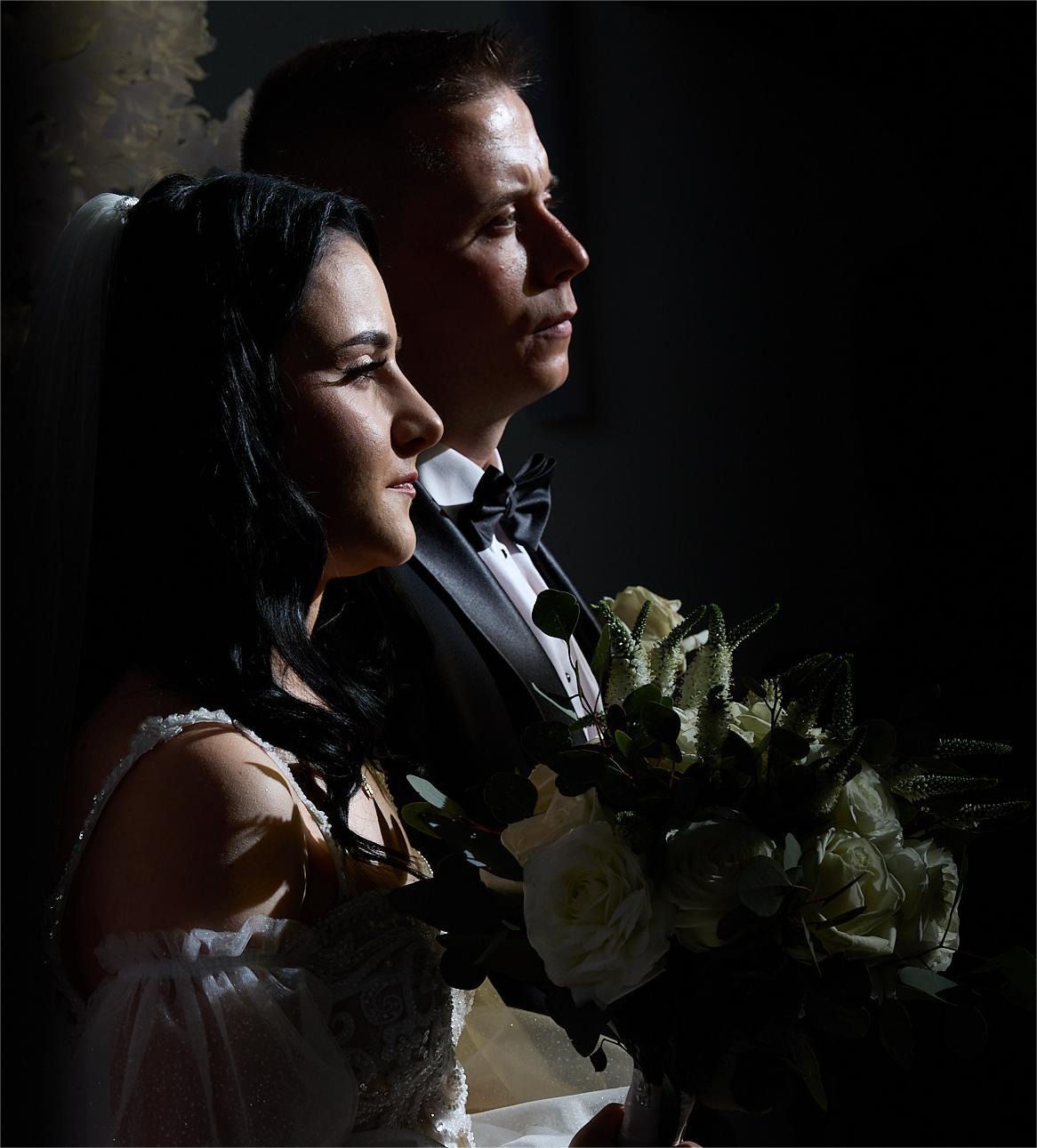 Elegancka para młoda w profilu, on w smokingu, ona w sukni z welonem, trzyma bukiet białych róż, oświetlona punktowo na ciemnym tle.