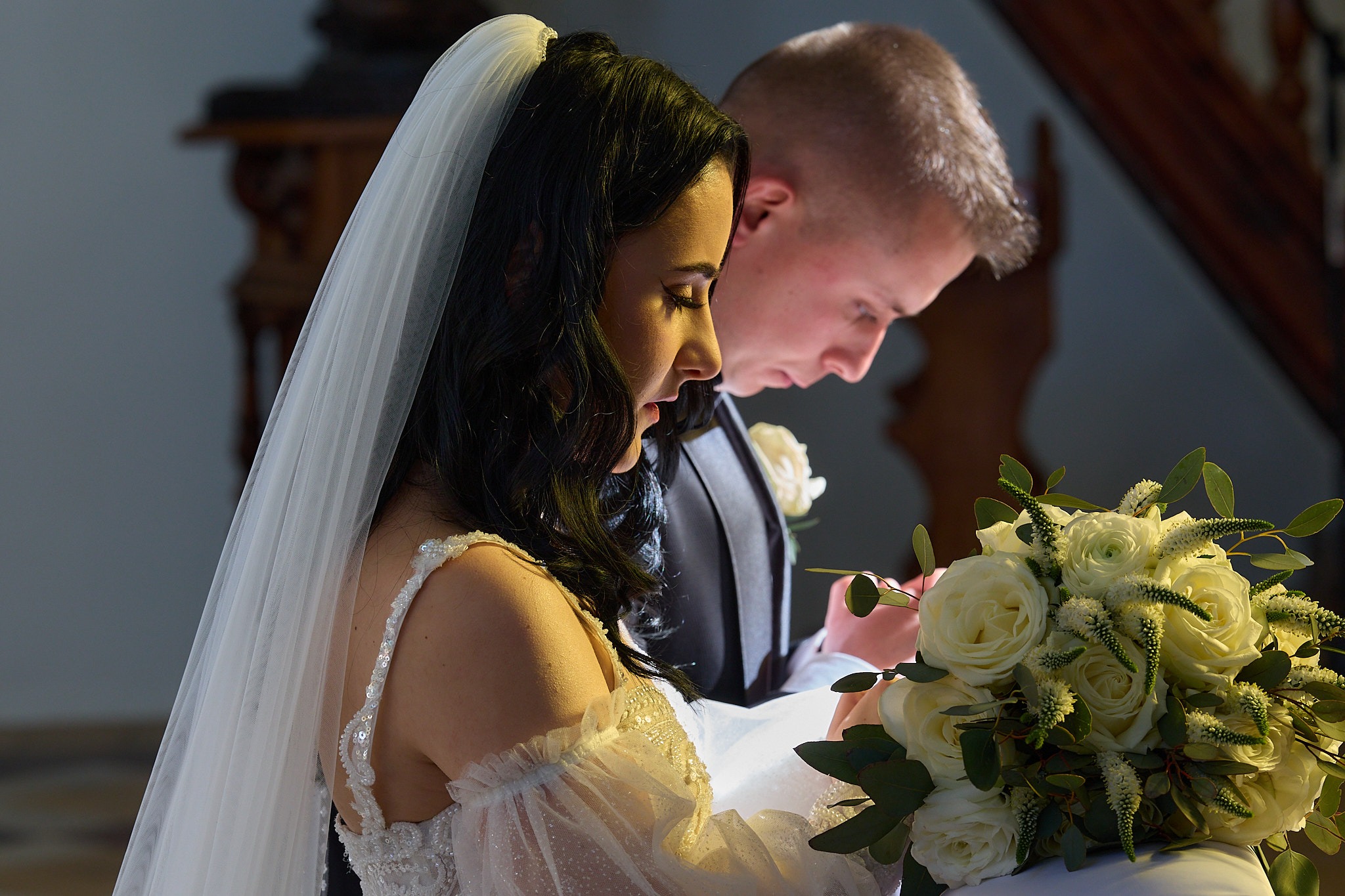 Profilowe ujęcie pary młodej podczas ceremonii ślubnej, z bukietem białych róż na pierwszym planie i delikatnym, rozproszonym oświetleniem.