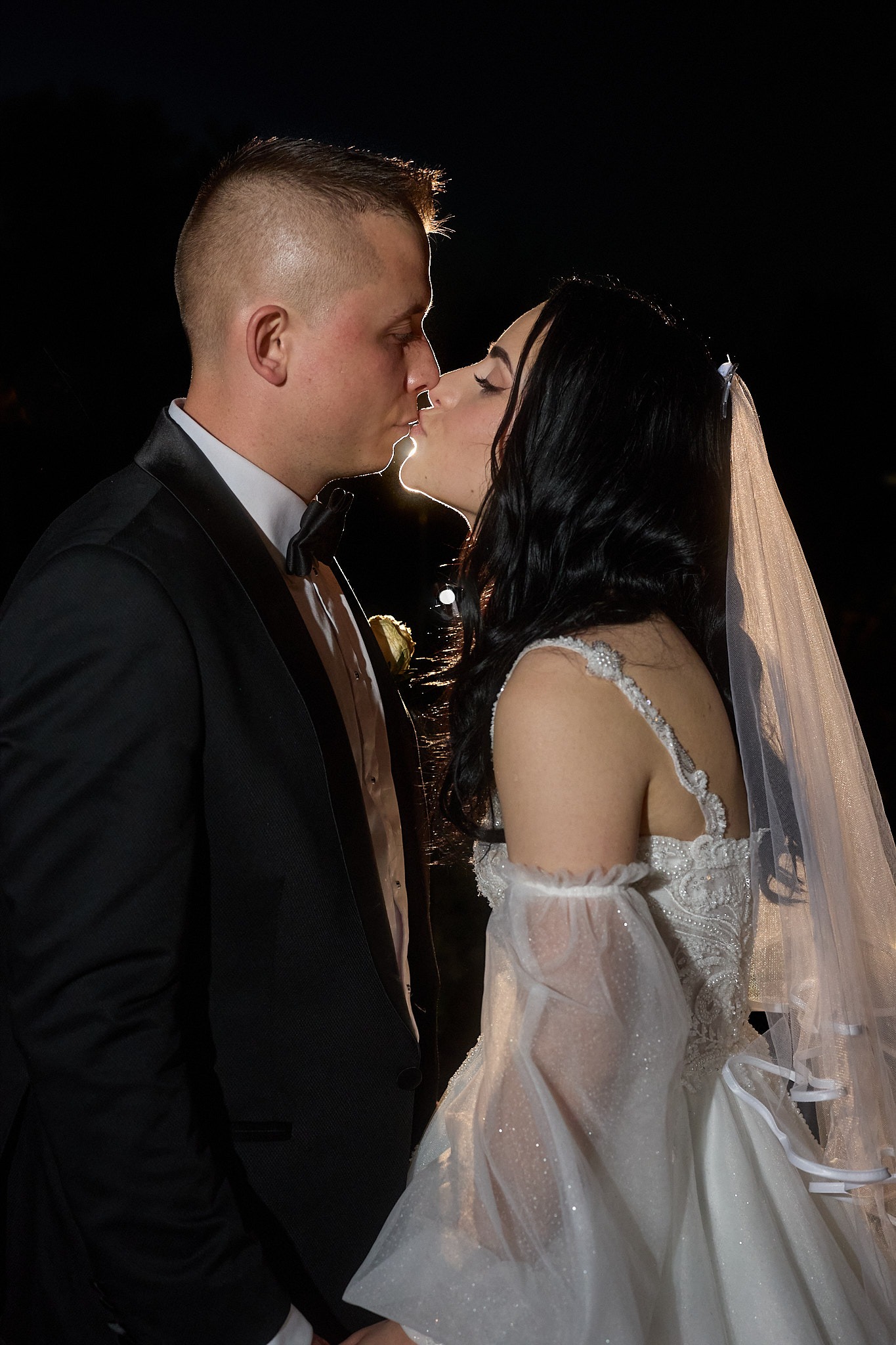 Para młoda w pocałunku na tle nocnego krajobrazu. Panna młoda w białej sukni z welonem, pan młody w czarnym smokingu. Romantyczna chwila uchwycona w ciemności.