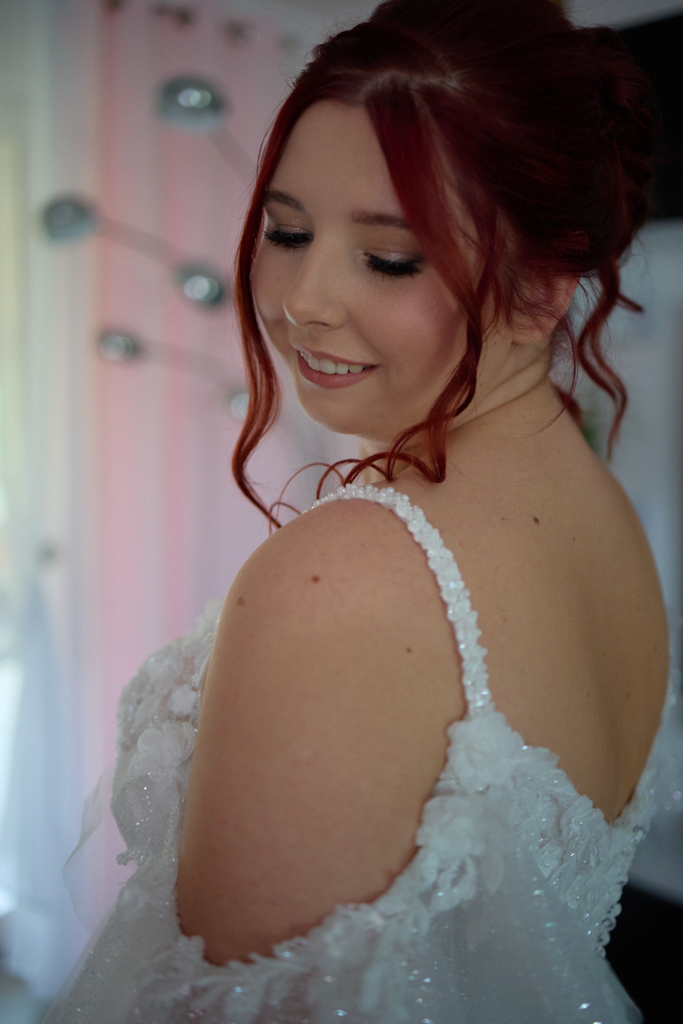 Uśmiechnięta panna młoda z rudymi włosami, w białej sukni z koronkowymi aplikacjami i zdobieniami na ramiączkach, patrzy w dół w delikatnym oświetleniu.