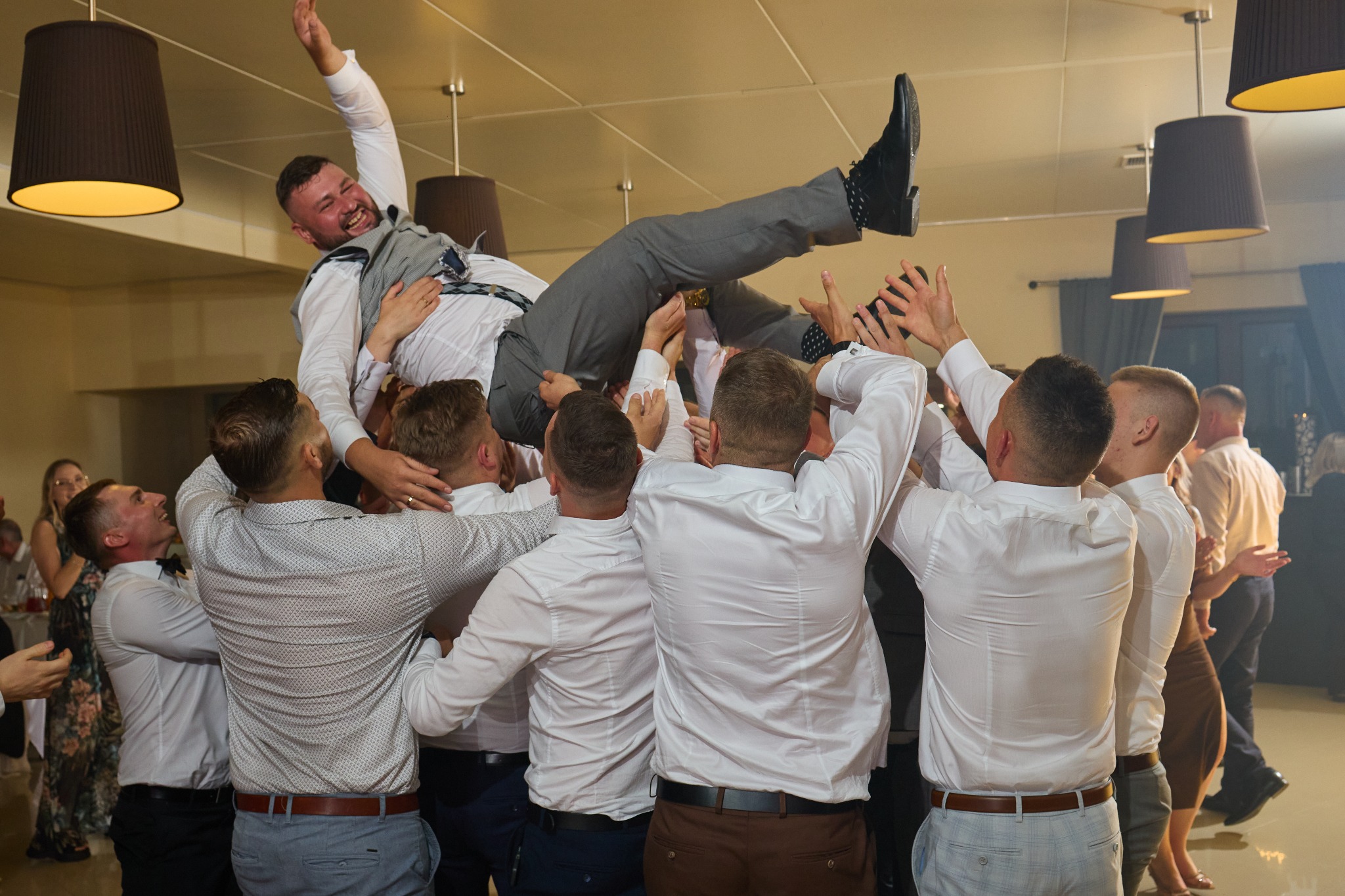 Radosny pan młody podrzucany przez gości weselnych w jasnym wnętrzu sali, widoczne lampy i fragmenty bawiących się osób.