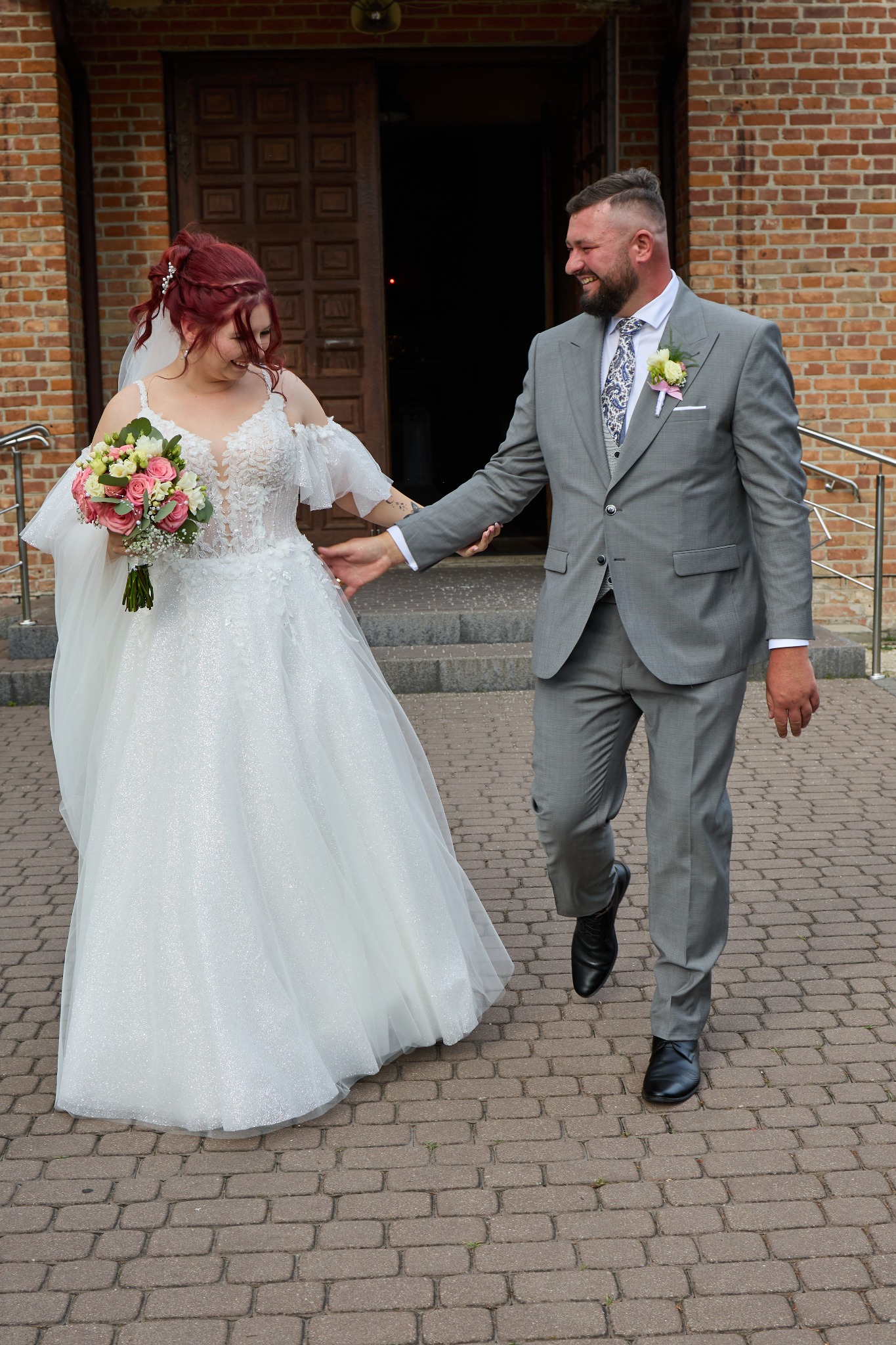 Para młoda wychodzi z kościoła. Panna młoda w białej sukni z bukietem, pan młody w szarym garniturze. Ceglany budynek w tle.