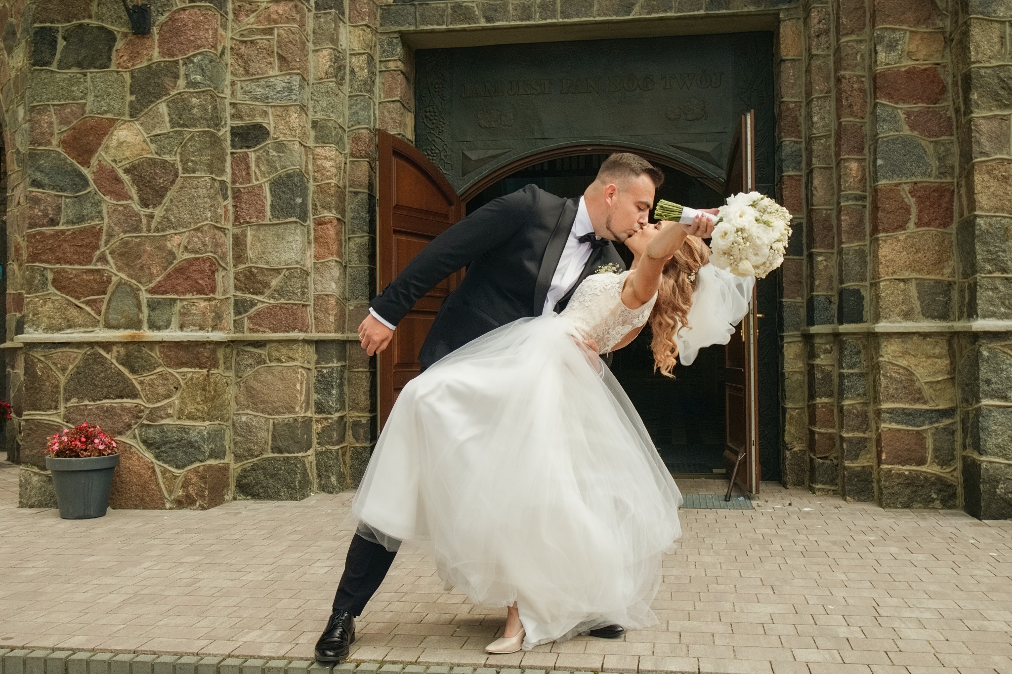Para młoda w tańcu przed kościołem o kamiennej fasadzie. Panna młoda w białej sukni, pan młody w czarnym smokingu. Bukiet w dłoniach panny młodej.