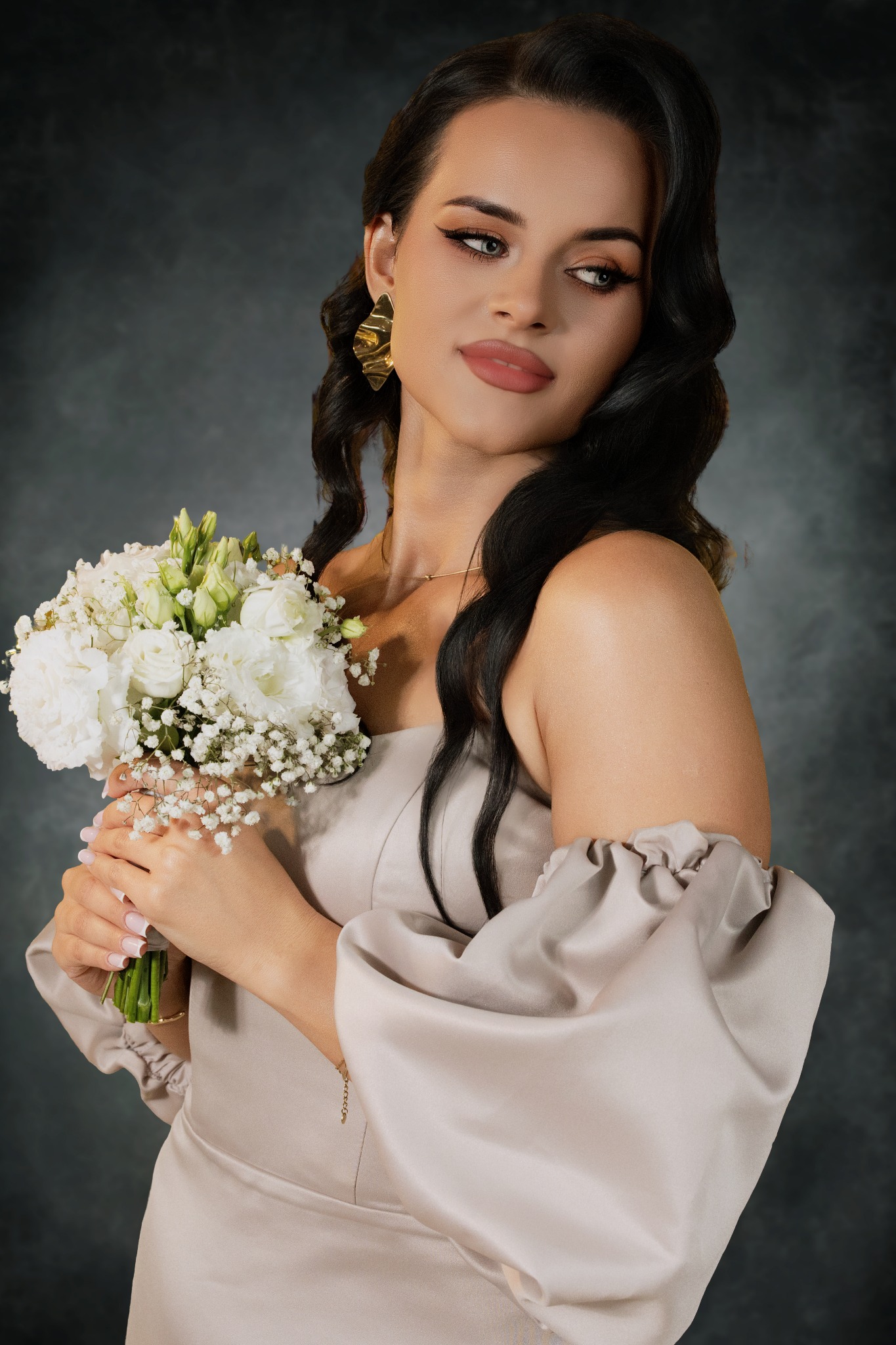 Elegancka kobieta w satynowej sukni z bukietem białych kwiatów, pozująca na tle ciemnego, teksturowanego tła. Widoczne złote kolczyki i subtelny makijaż.