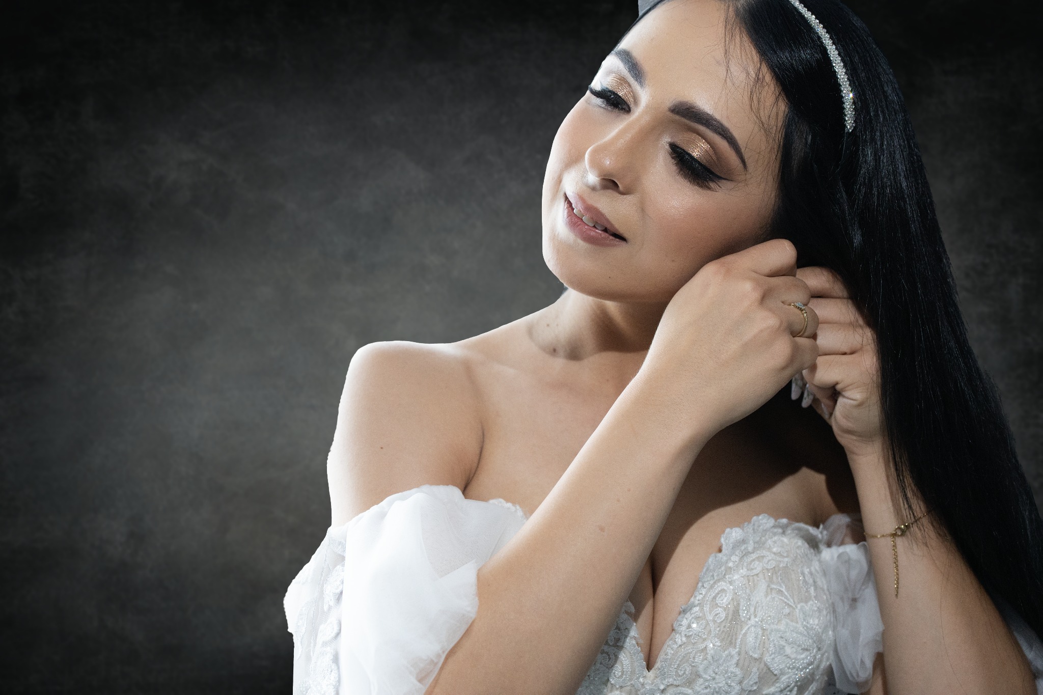 Panna młoda w sukni ślubnej, z diademem we włosach, zakłada kolczyk. Makijaż z błyszczącym cieniem. Tło ciemne, lekko rozmyte.