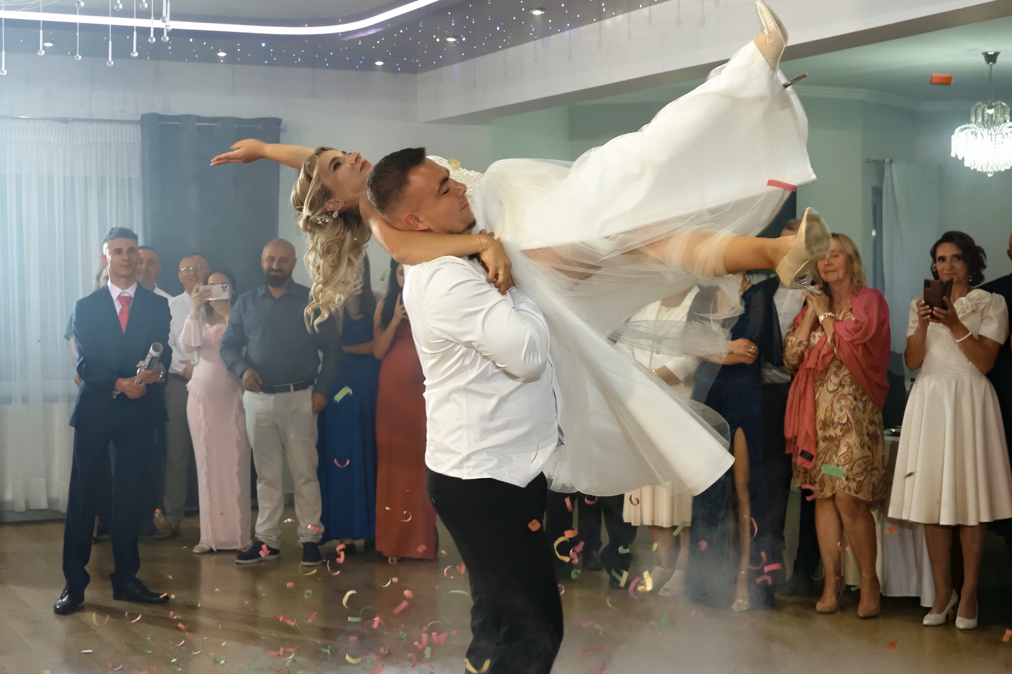 Pan młody trzyma uniesioną pannę młodą na parkiecie, w tle goście z telefonami, konfetti na podłodze. Radosna atmosfera wesela.