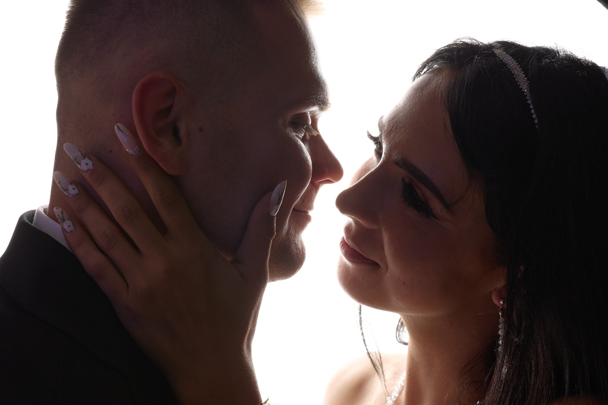 Silhouetted close-up of a bride with a rhinestone headband and floral-painted nails gently touching the groom's face, backlight emphasizing their profiles.