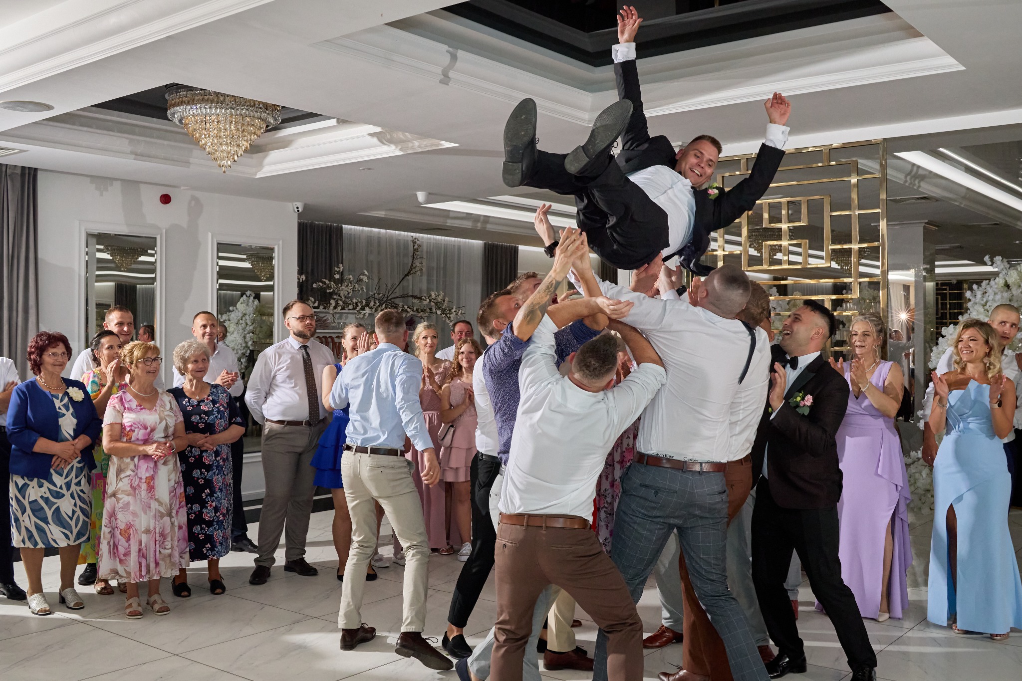 Uśmiechnięty pan młody podrzucany w górę przez gości weselnych, w tle elegancki wystrój sali z kryształowym żyrandolem i bawiący się goście.