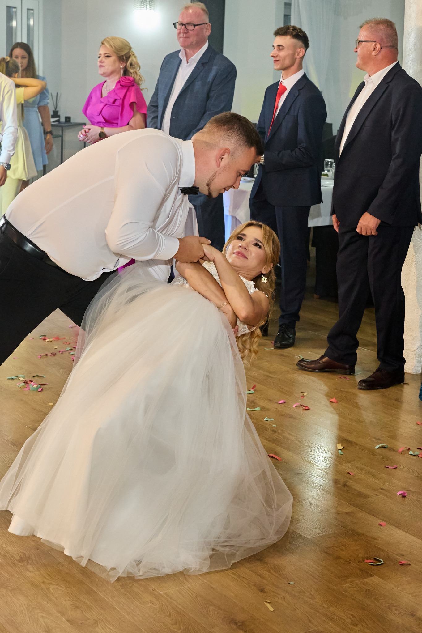 Para młoda tańczy taniec weselny, on w białej koszuli i muszce, ona w białej sukni, w tle goście weselni i konfetti na podłodze.