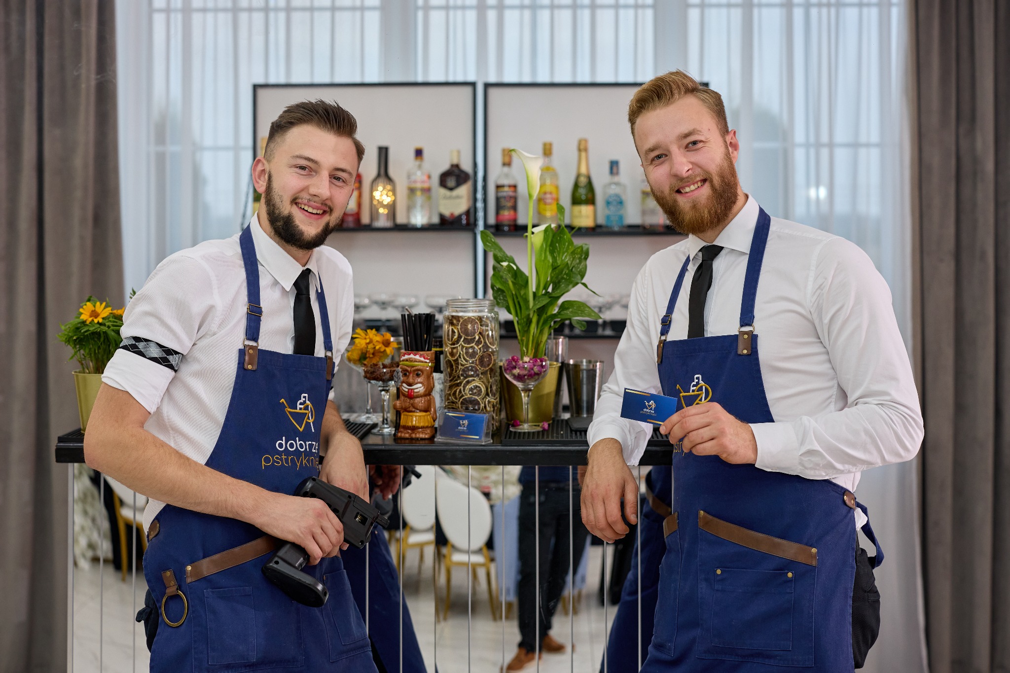 Dwóch uśmiechniętych barmanów w granatowych fartuchach z logo firmy, za barem z dekoracjami i butelkami alkoholu. Jeden trzyma metkownicę, drugi wizytówkę.