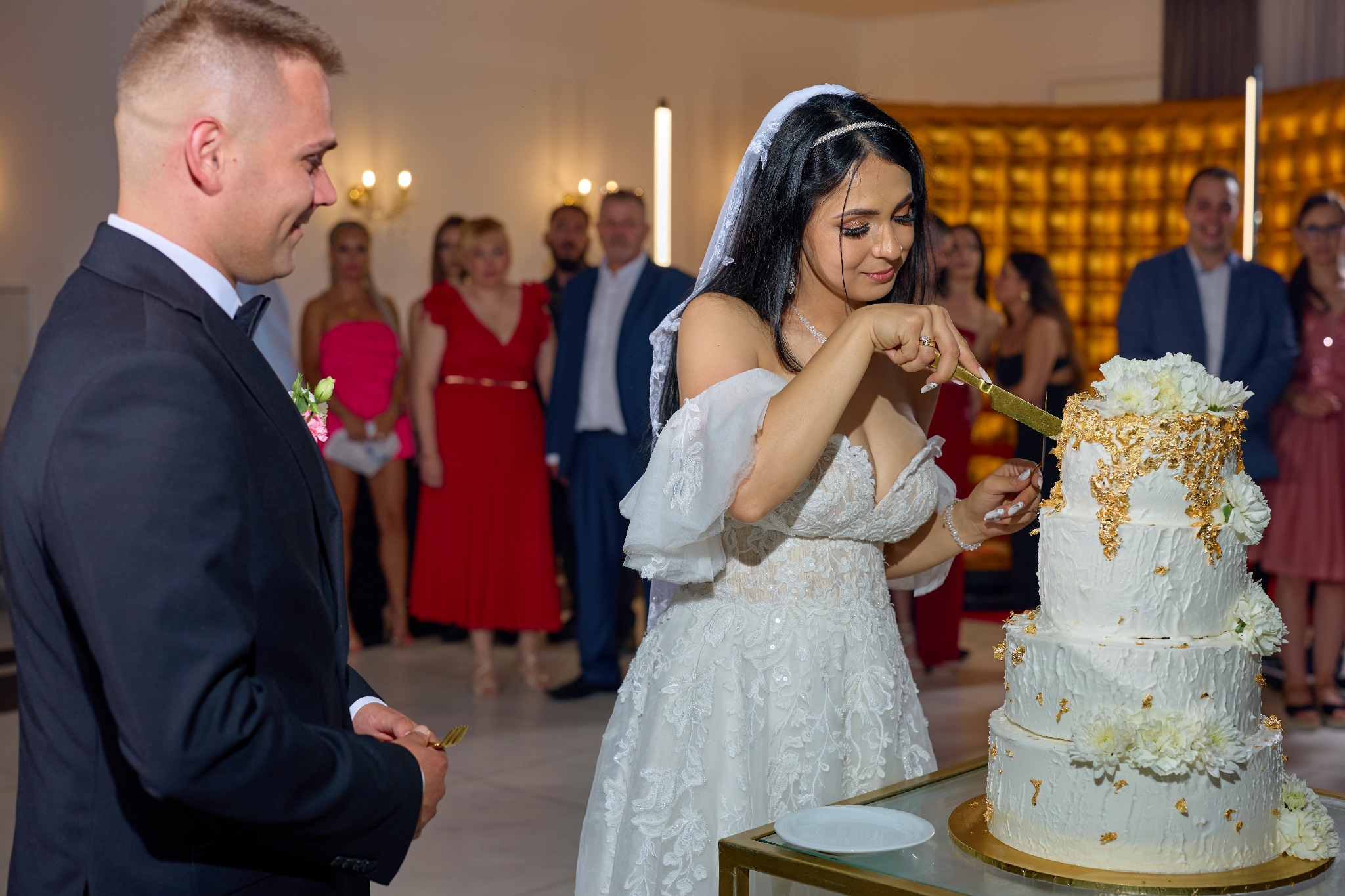 Uśmiechnięta panna młoda w białej sukni kroi tort weselny zdobiony złotem, obok stoi pan młody. W tle goście weselni i dekoracje sali.