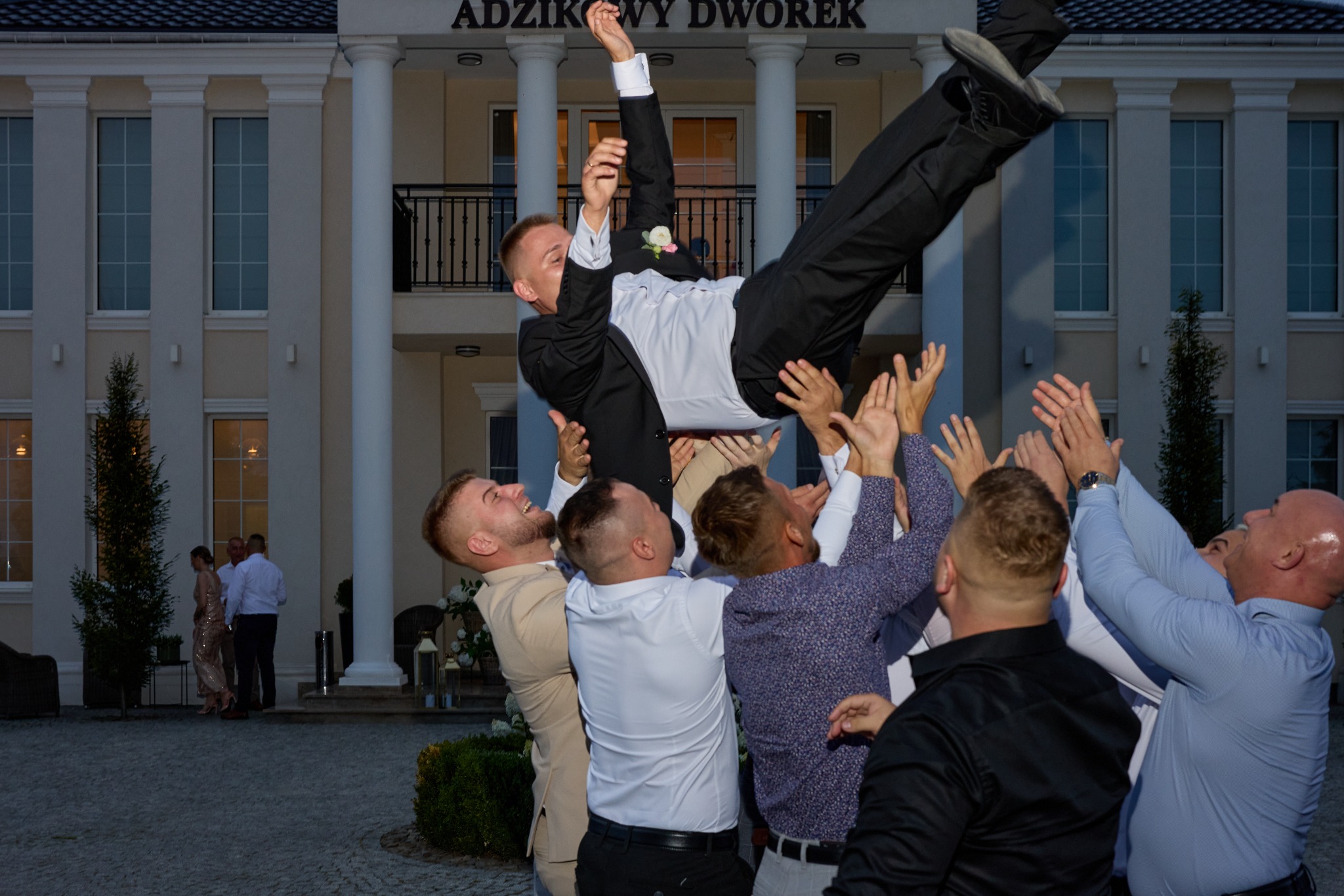 Radosne podrzucanie pana młodego przez gości weselnych przed budynkiem 'Adzikowy Dworek' w dniu ślubu. W tle para w eleganckich strojach.