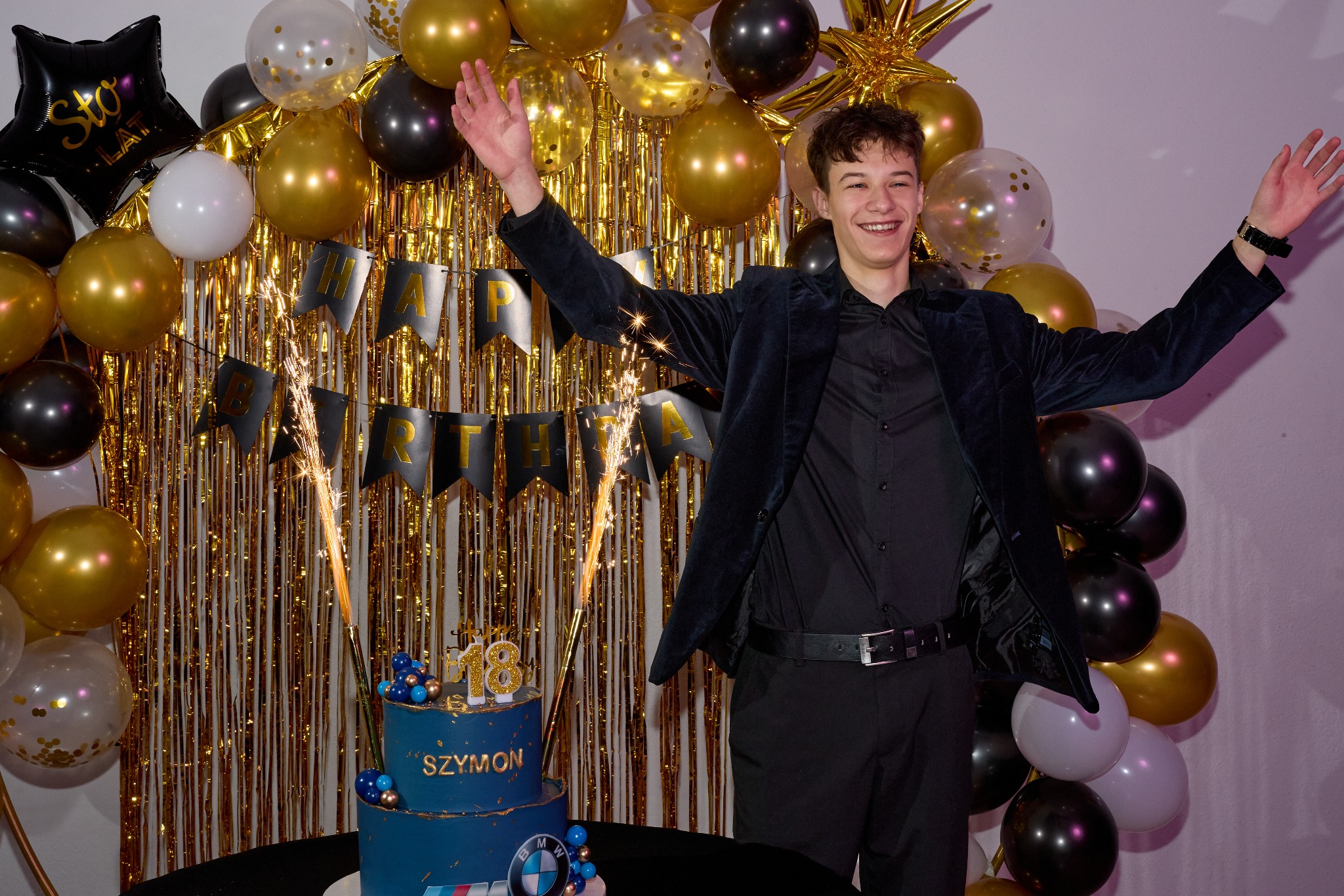Uśmiechnięty nastolatek w czarnym stroju, z rękami w górze, świętuje 18 urodziny na tle złotych dekoracji, tortu z fajerwerkami i balonów.
