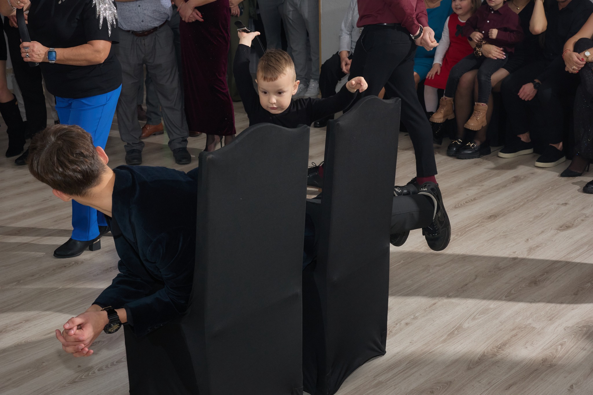 Chłopiec w czarnym stroju wyłania się zza dwóch krzeseł, podczas gdy fotograf w aksamitnej marynarce leży na podłodze, próbując uchwycić unikalne ujęcie podczas uroczystości.