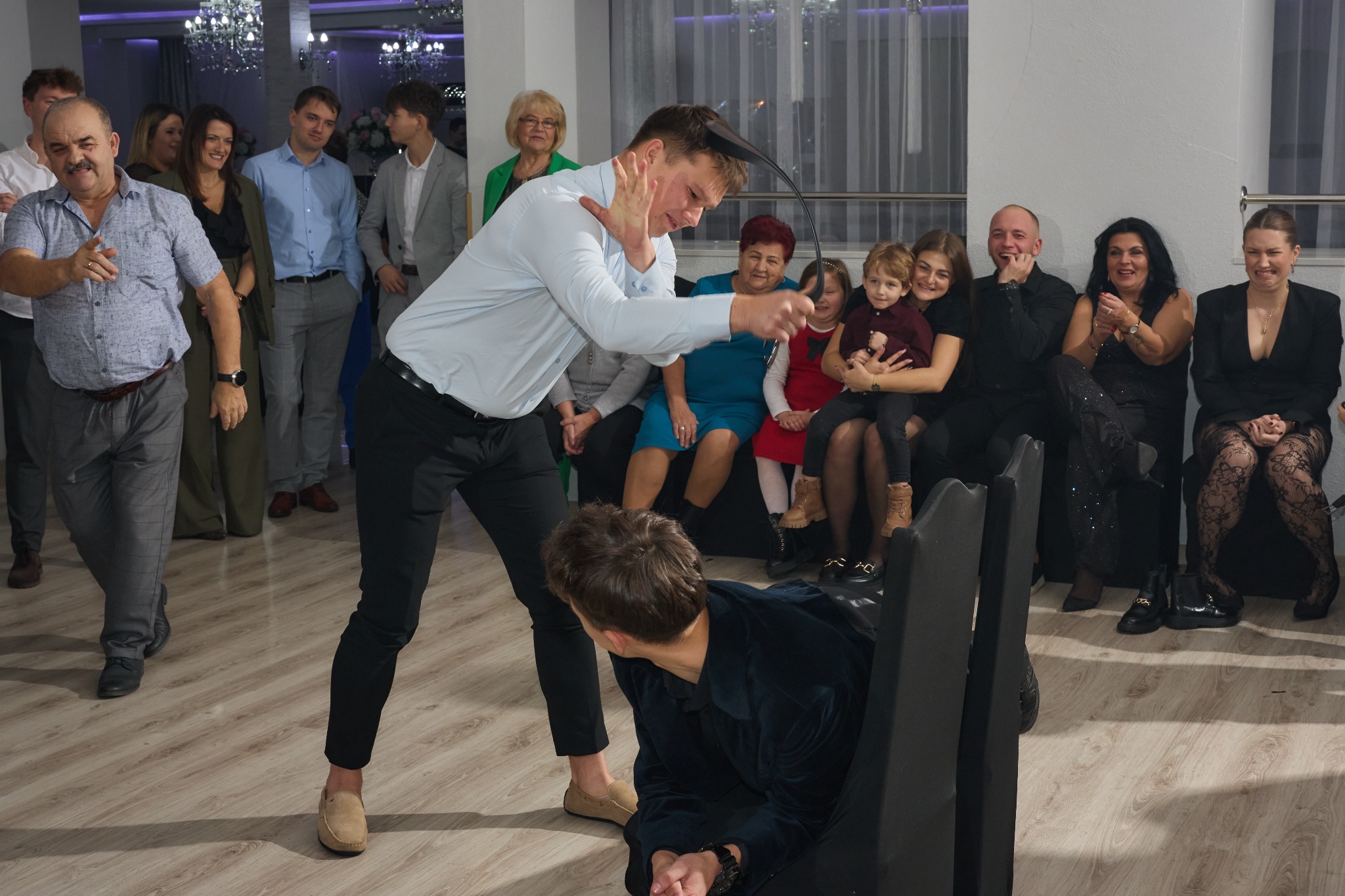 Dynamiczne ujęcie z oczepin na weselu: mężczyzna w koszuli łapie welon, drugi klęczy. Goście weselni obserwują z zaciekawieniem, w tle kryształowe żyrandole.