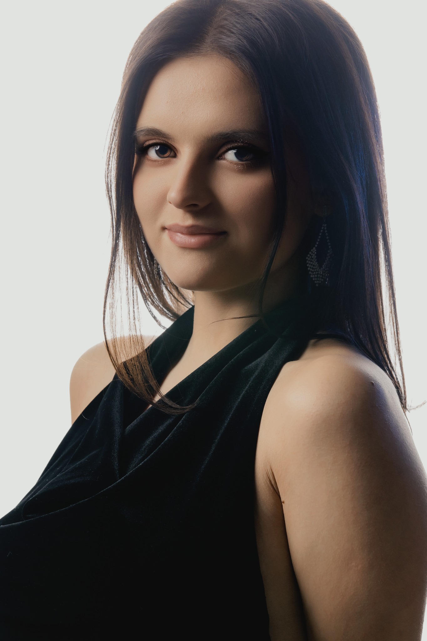 Portret młodej kobiety z ciemnymi włosami, ubrana w czarną sukienkę, z delikatnym uśmiechem i błyszczącymi kolczykami na jasnym tle. Subtelny makijaż podkreśla jej urodę.