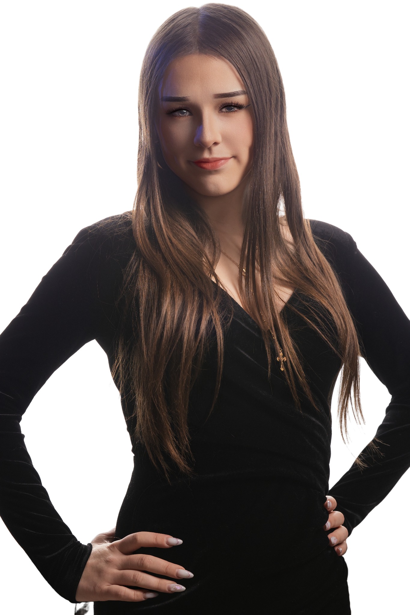 Portret młodej kobiety w czarnej sukience na białym tle, z rękami na biodrach i delikatnym uśmiechem. Widoczny złoty łańcuszek z krzyżykiem.