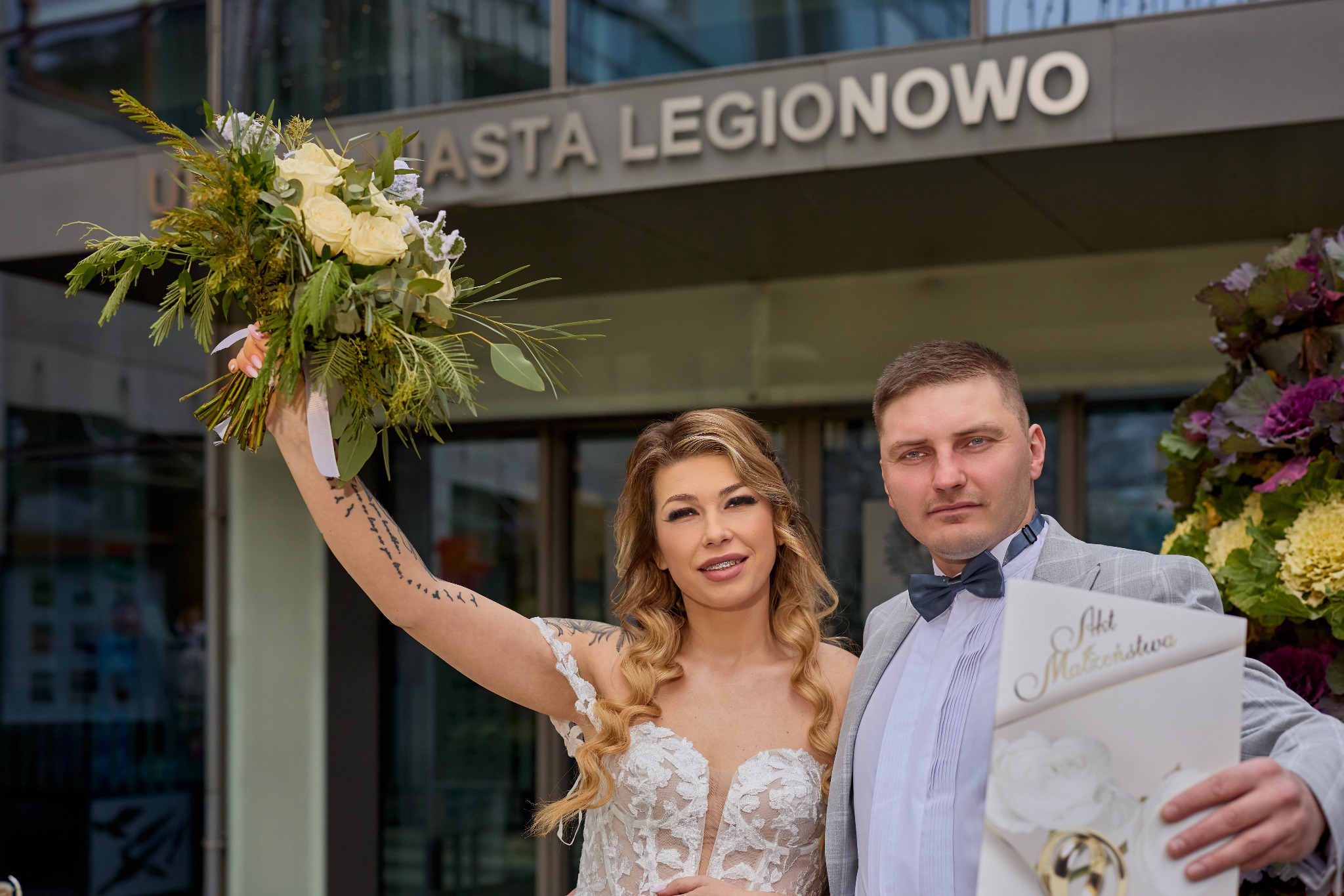 Para młoda pozuje z bukietem i aktem małżeństwa na tle budynku z napisem MIASTA LEGIONOWO. Panna młoda z tatuażem na ramieniu.