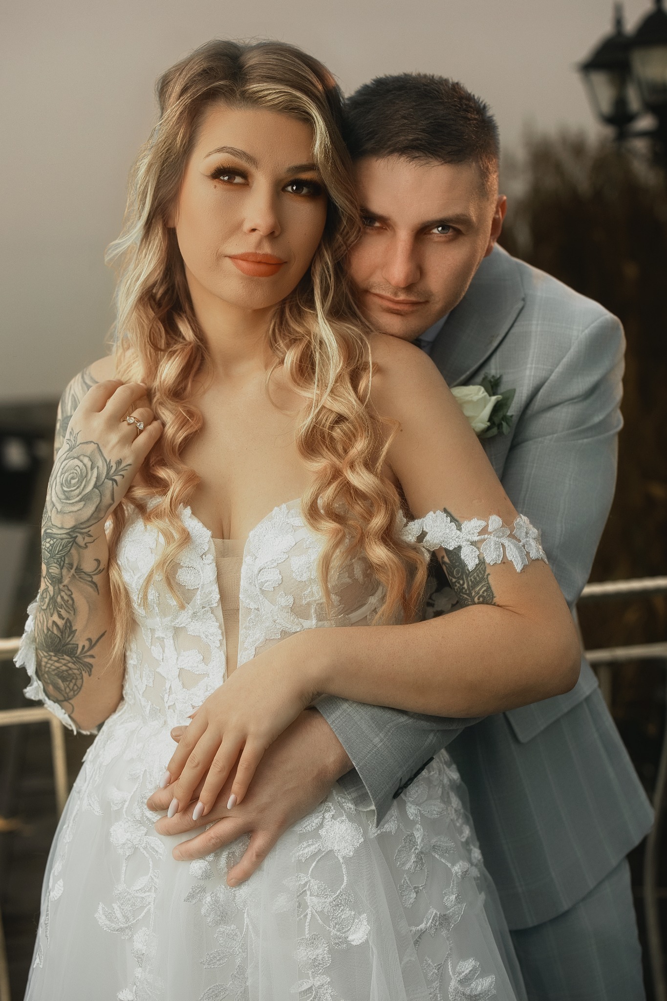 Uśmiechnięta para młoda w objęciach na tle balkonu. Panna młoda w koronkowej sukni z tatuażami, pan młody w szarym garniturze z butonierką. Romantyczna sesja ślubna.