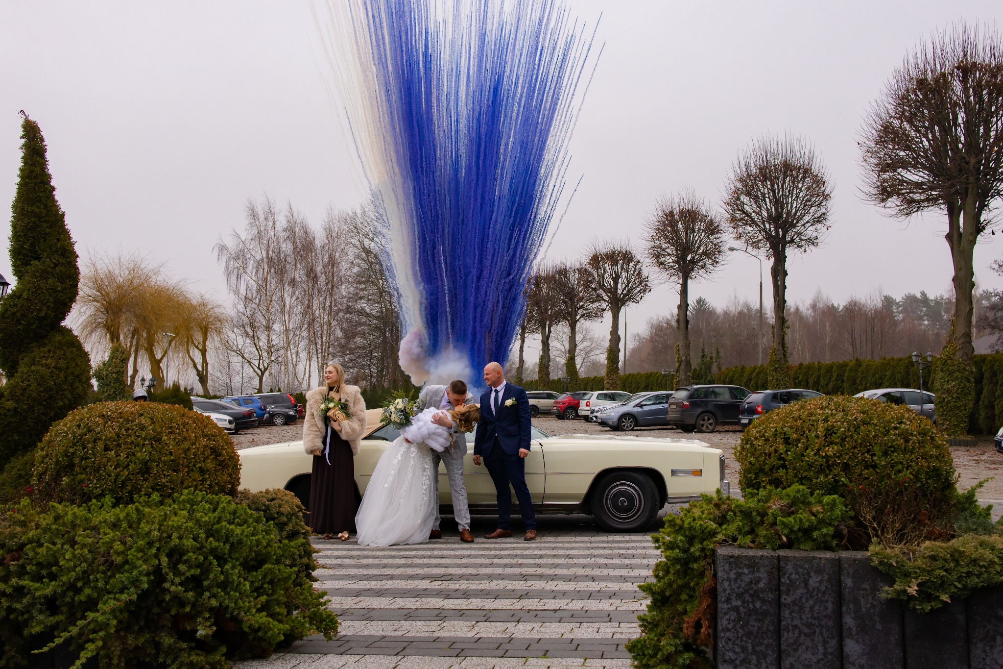 Para młoda w objęciach na tle białego samochodu Cadillac, z niebieskimi i białymi fajerwerkami w tle. Goście weselni i parking w tle. Ujęcie z zewnątrz.