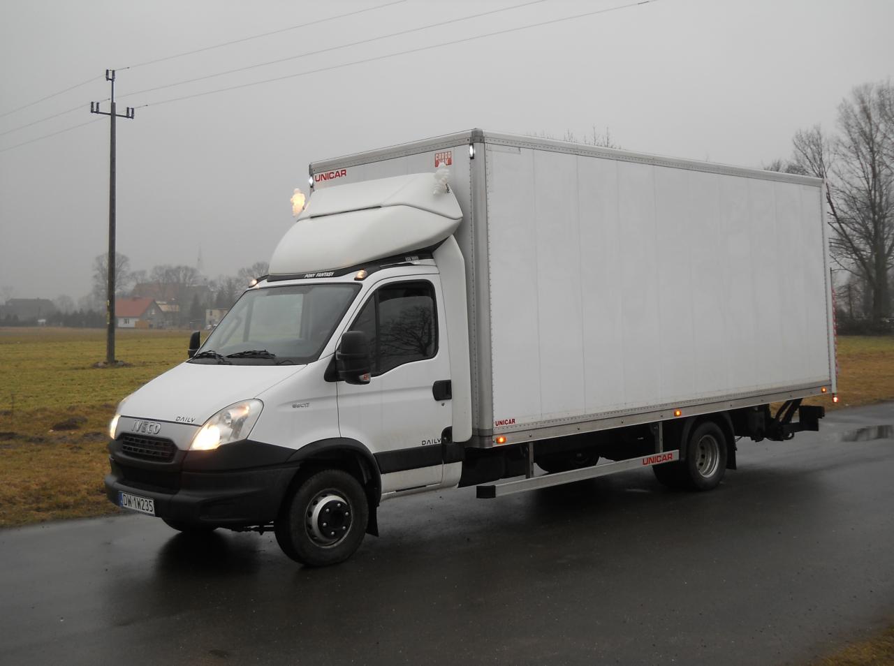 Biały samochód dostawczy Iveco Daily z zabudową kontenerową Unicar, zaparkowany na mokrym asfalcie w pochmurny dzień, widoczny numer rejestracyjny DW 1W235.