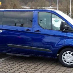 Niebieski bus marki Ford Transit z przyciemnianymi szybami, zaparkowany na parkingu w zimowej scenerii.