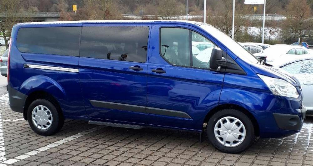 Niebieski bus marki Ford Transit z przyciemnianymi szybami, zaparkowany na parkingu w zimowej scenerii.