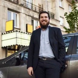 Uśmiechnięty mężczyzna w garniturze stoi obok ciemnoszarego samochodu na tle budynku z oświetlonym wejściem.