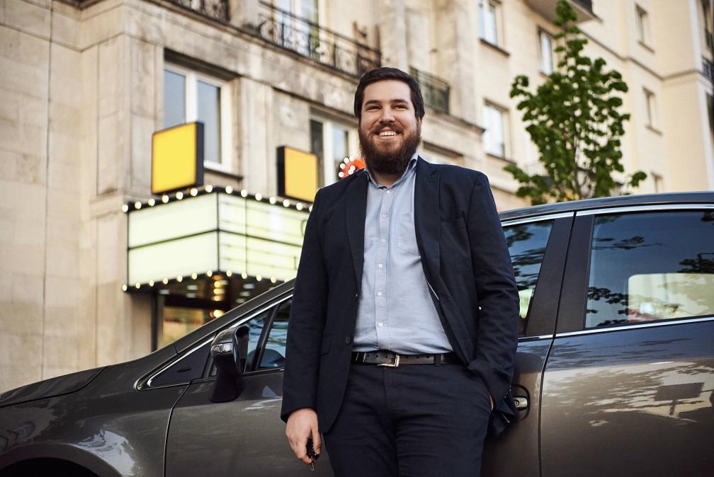 Uśmiechnięty mężczyzna w garniturze stoi obok ciemnoszarego samochodu na tle budynku z oświetlonym wejściem.
