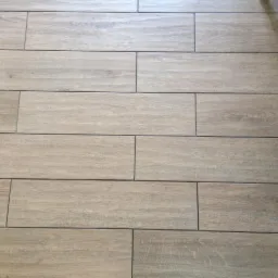 Ułożona podłoga z płytek imitujących drewno w odcieniach beżu i brązu, widoczne fugi i fragment nogi krzesła z boku.